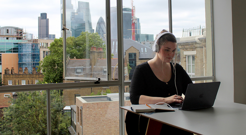 Image of a person working on a laptop, with a city skyline visible in the background through a window.